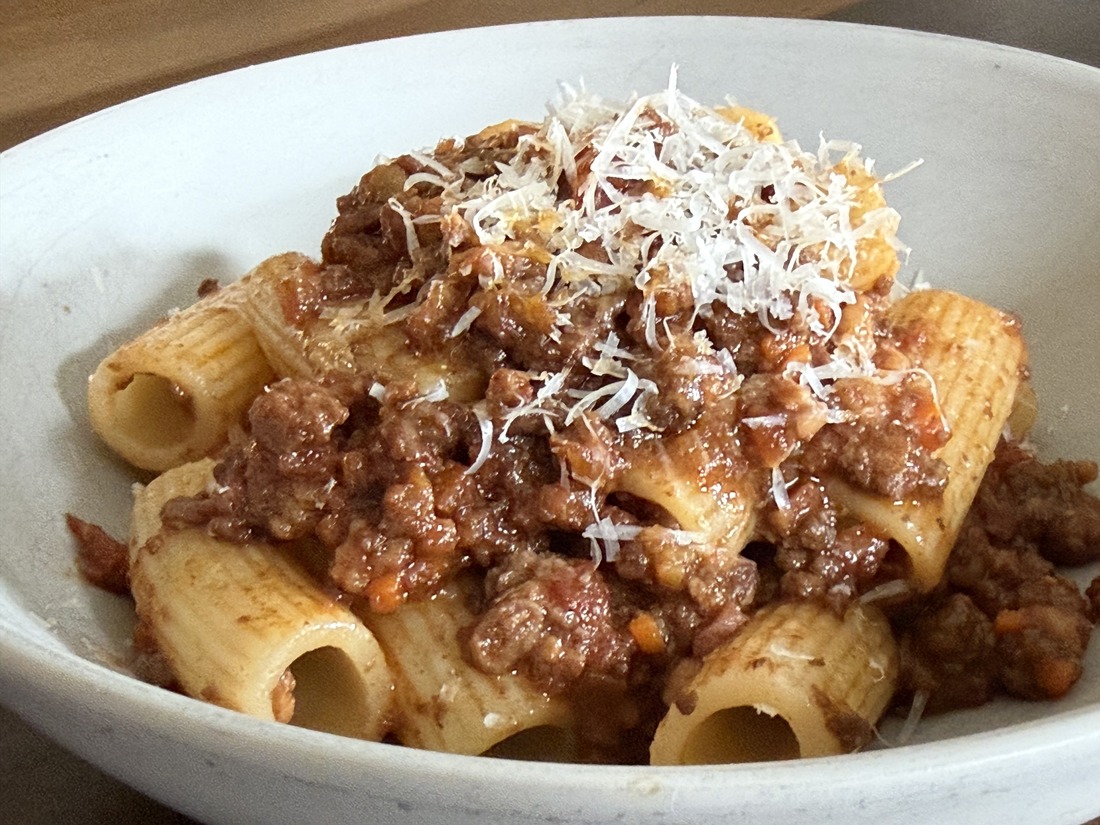 Rigatoni in rich bolognese, piled in a white bowl under a heavy shower of grated Parmesan