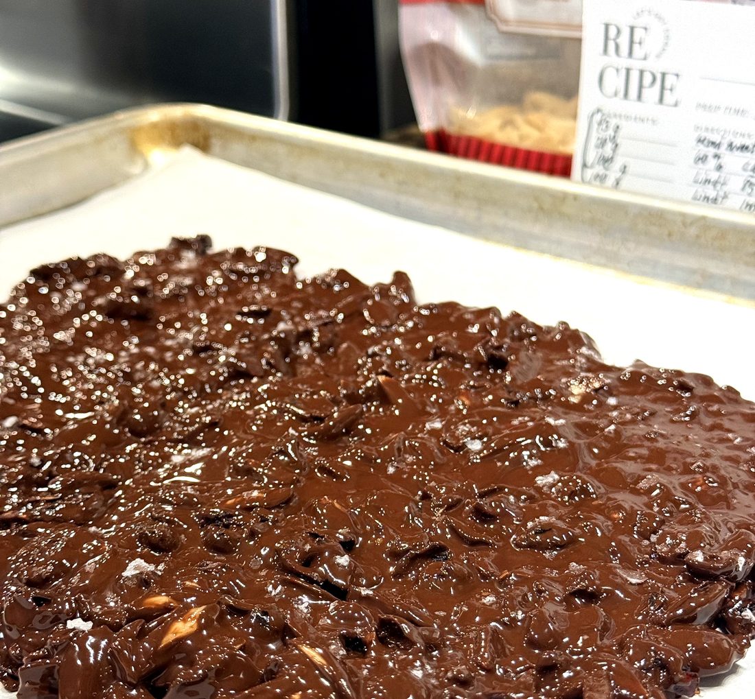 Dark chocolate bark with toasted almonds and cranberries spread on parchment, still glossy and warm before setting