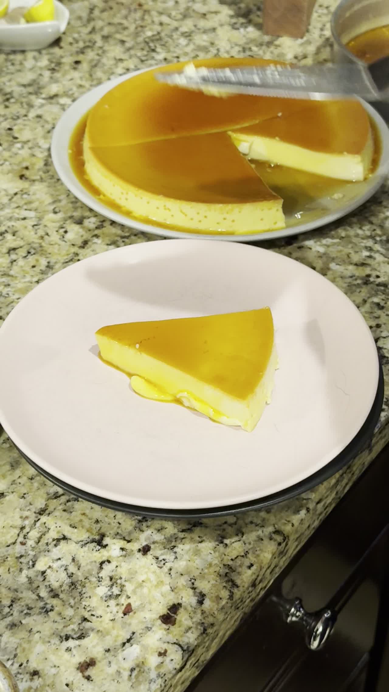 A slice of flan plated on a pink plate, whole flan with cascading caramel in the background
