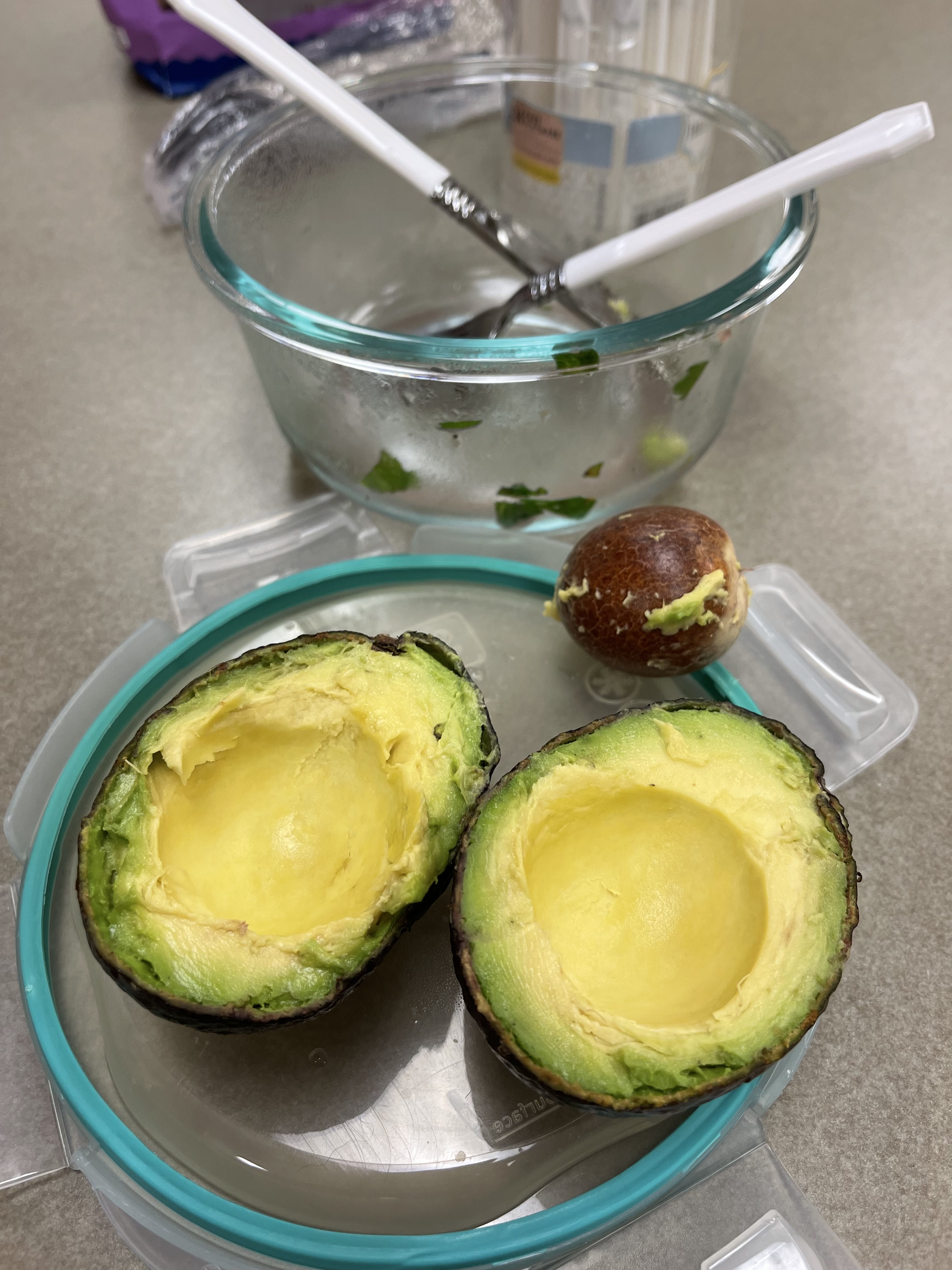 Halved avocado with pit removed, resting on a teal container lid beside a glass mixing bowl