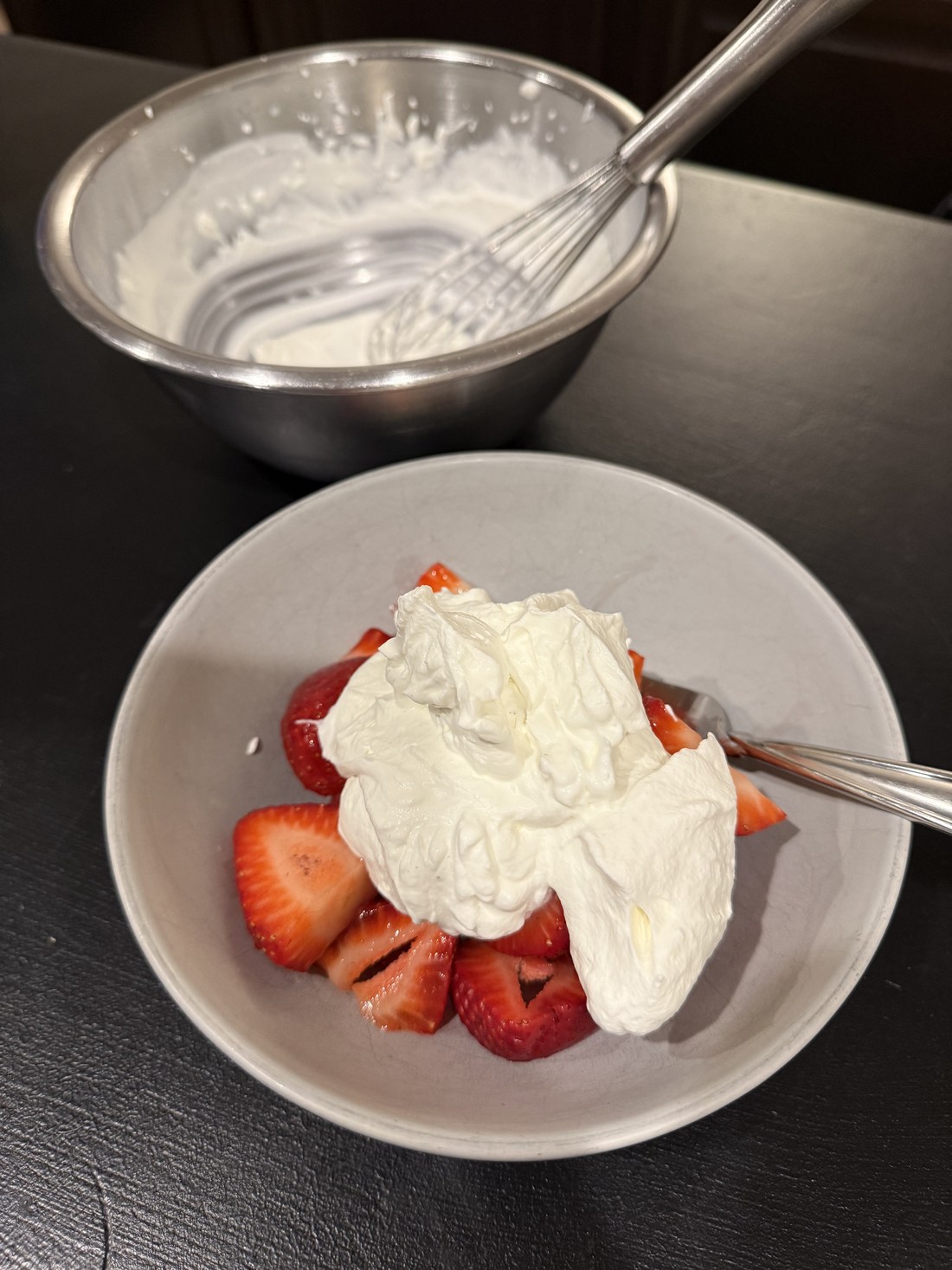 Sliced macerated strawberries in their syrup under a mound of hand-whipped cream, with the mixing bowl and whisk still on the counter behind