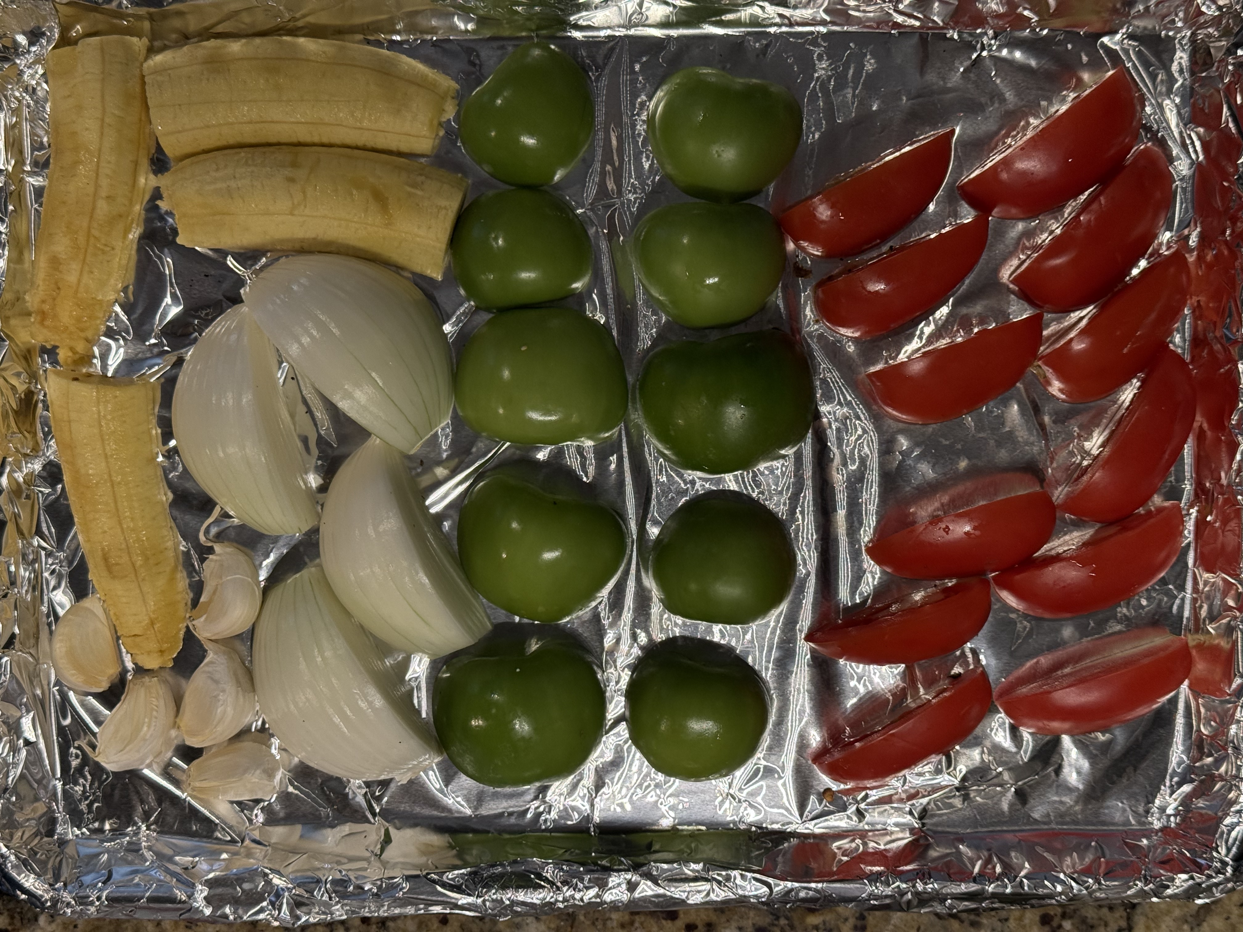 Plantain, onion, garlic, tomatillos, and tomatoes arranged on a foil-lined sheet pan, ready to broil