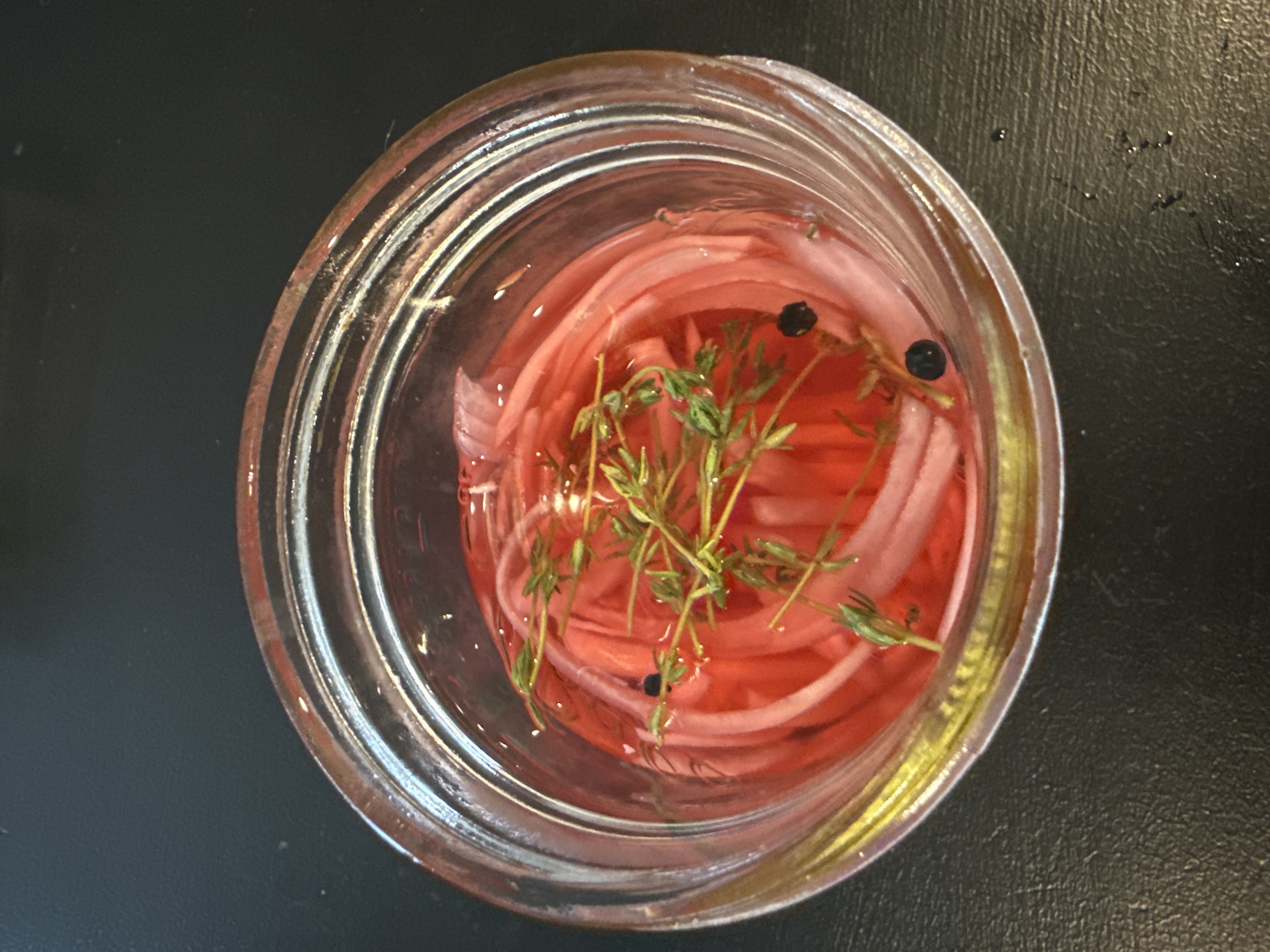 Pickled red onions in a mason jar viewed from above — vivid pink brine with thyme sprigs and whole black peppercorns