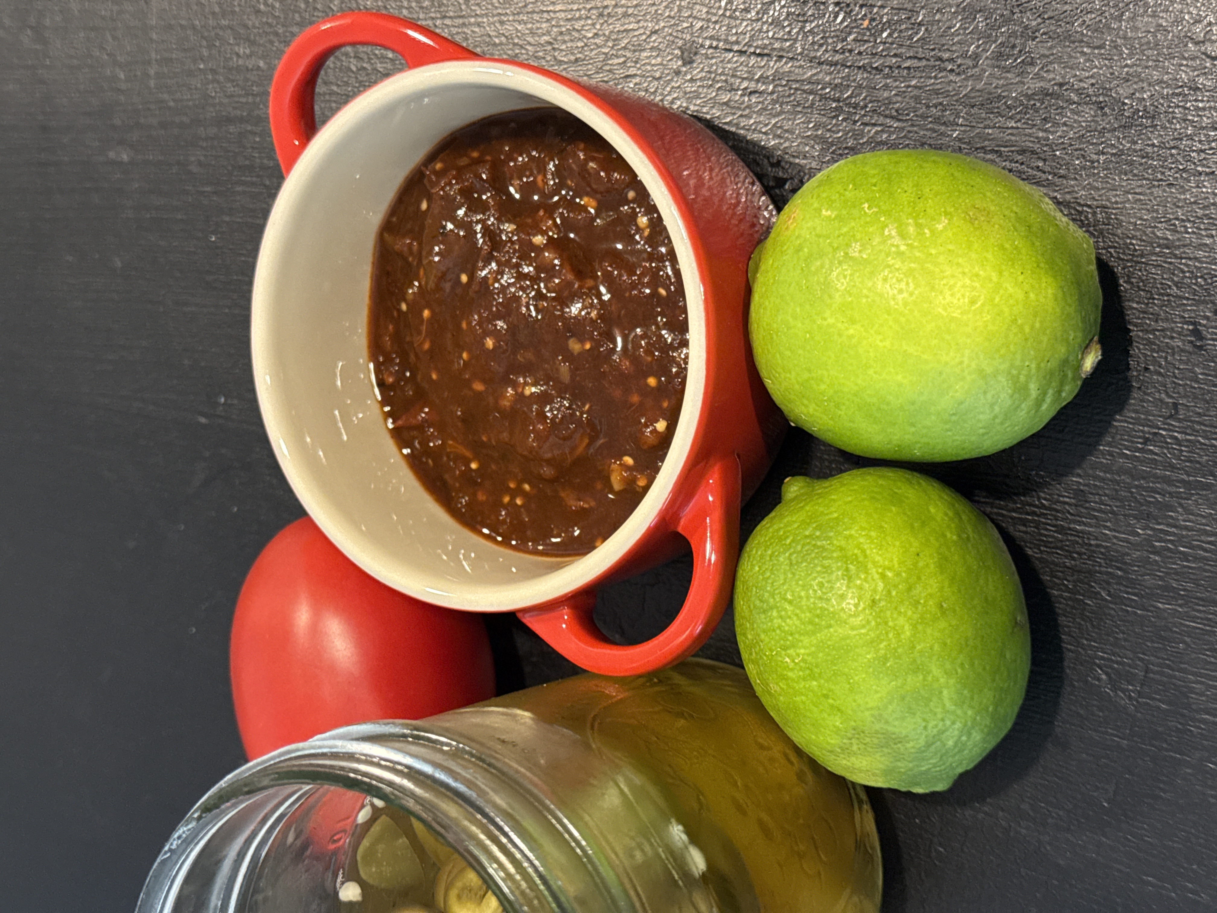 Deep dark roasted chile salsa in a red crock, styled on a slate board with fresh limes and a tomato