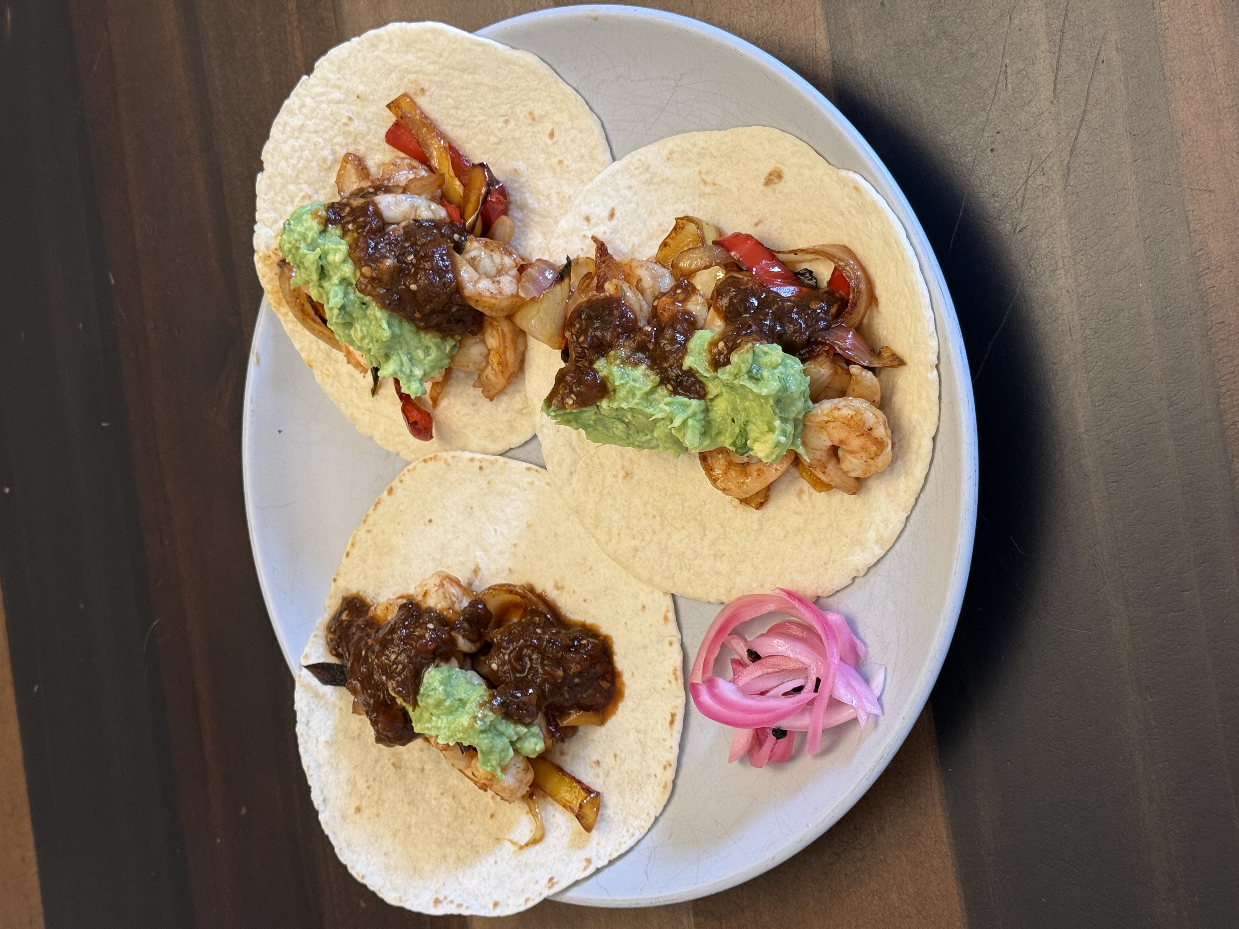 Three spicy shrimp fajita tacos on flour tortillas with guacamole and dark marinade glaze, pickled red onions on the side