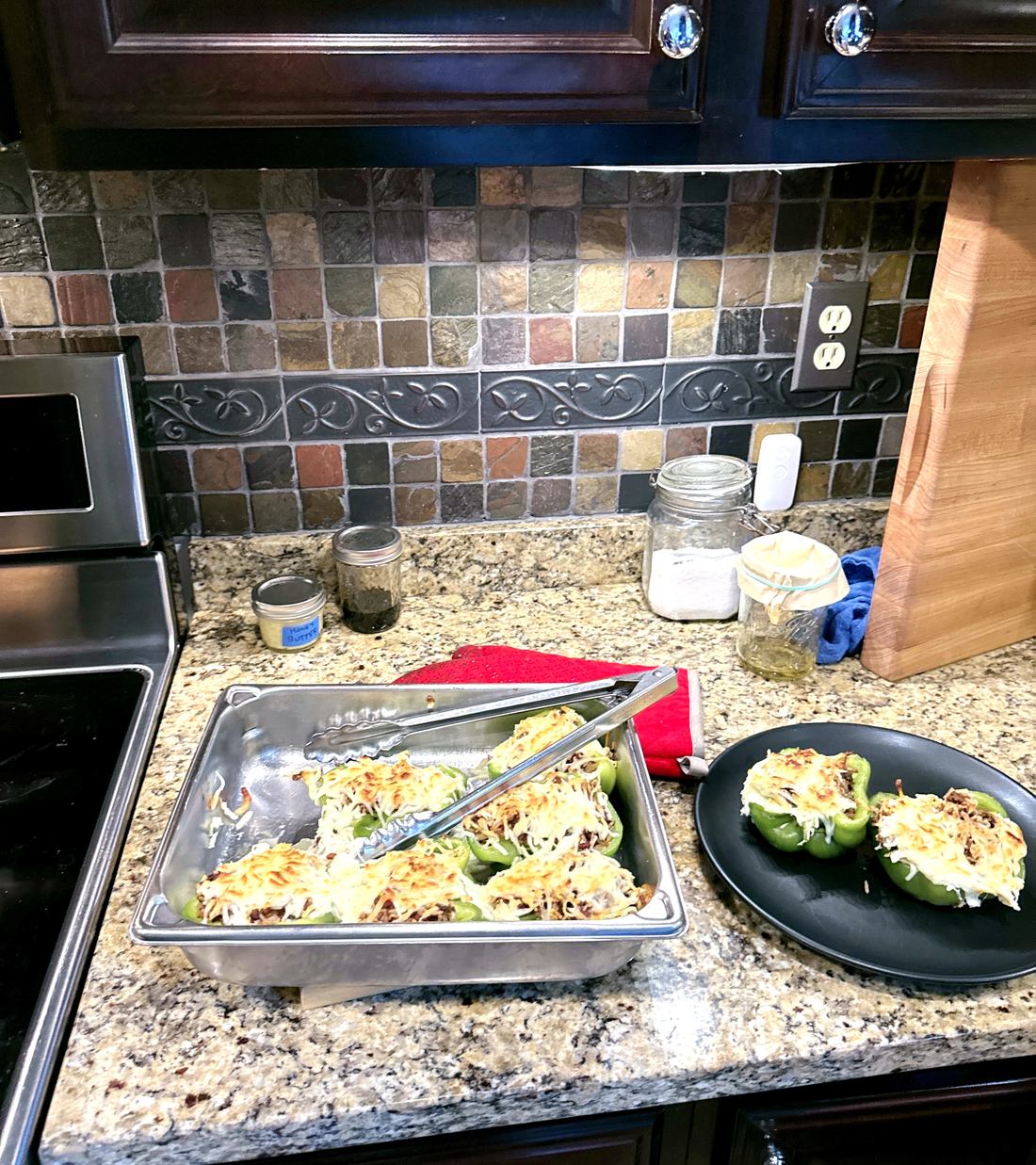 A hotel pan of baked stuffed peppers on the counter with tongs, next to a plated serving