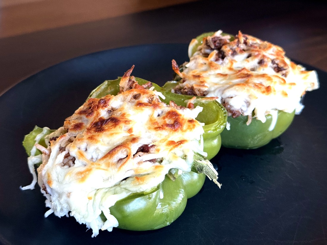 Two bolognese-stuffed pepper halves in a dark bowl, topped with browned and bubbling mozzarella