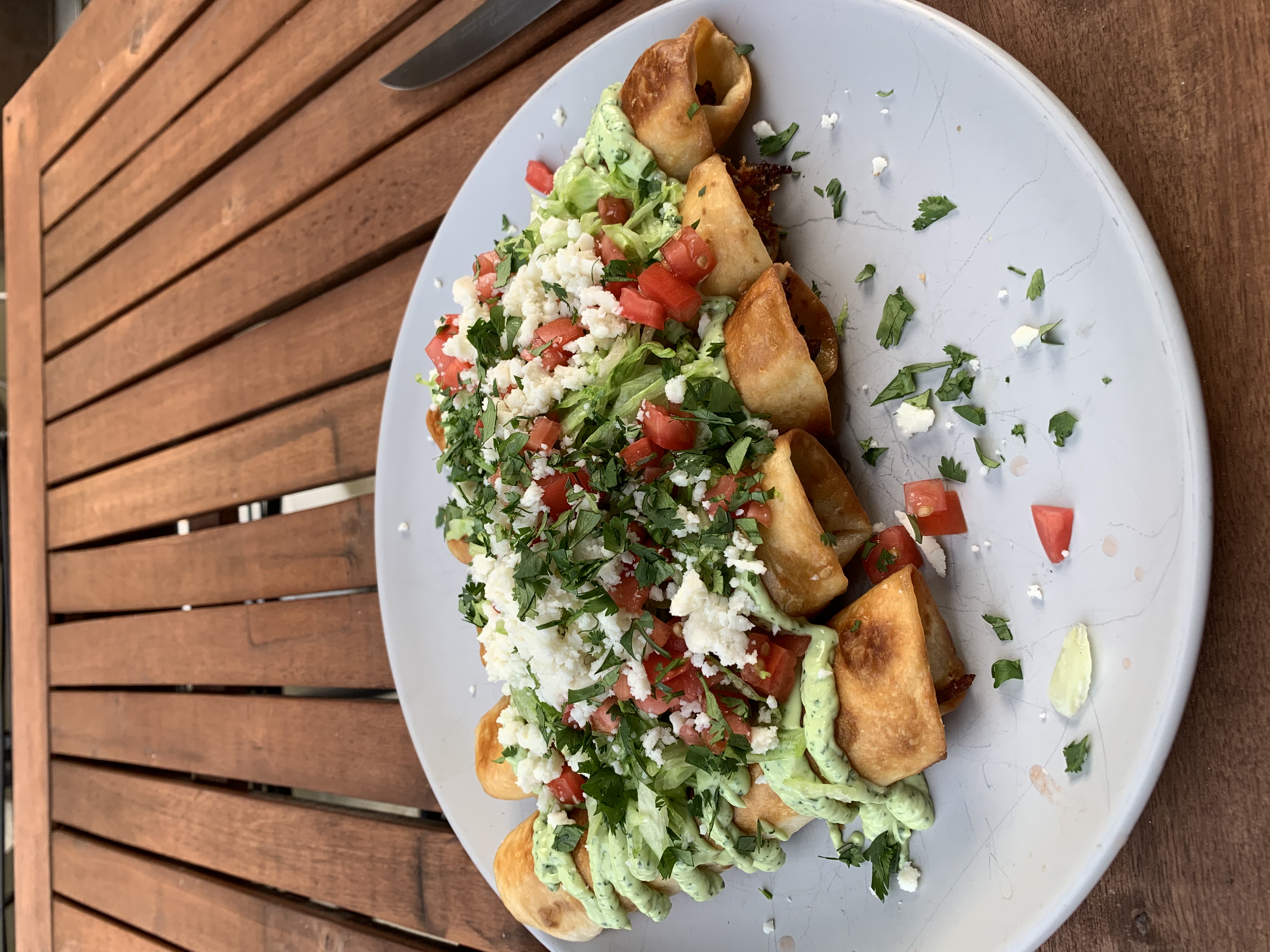 Crispy rolled taquitos on a white oval plate, topped with cilantro crema, shredded lettuce, pico de gallo, queso fresco, and chopped cilantro