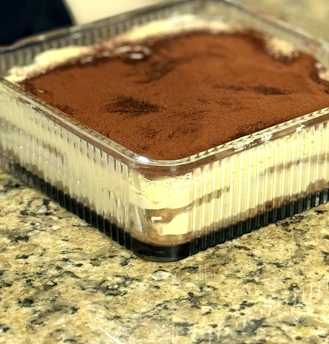 Tiramisu in a ribbed glass dish, viewed from an angle showing the cocoa-dusted top and layered sides