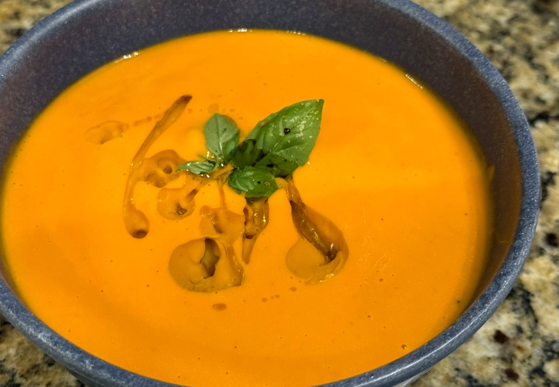 A bowl of smooth orange tomato and white bean soup, swirled with olive oil and topped with a sprig of fresh basil