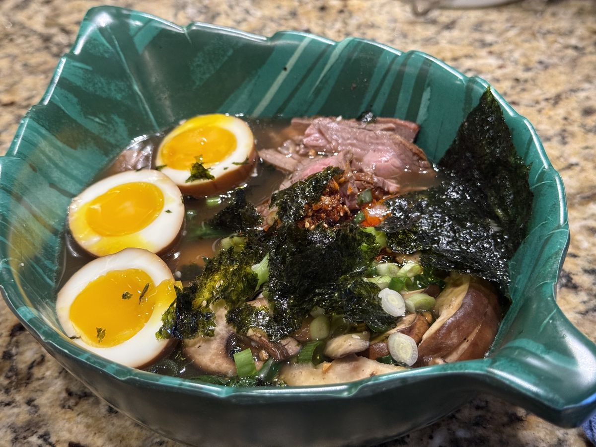 Asian noodle bowl — sliced seared flank steak, three jammy soy-marinated egg halves, shiitake mushrooms, nori, and scallions in a dark sesame broth in a teal bowl